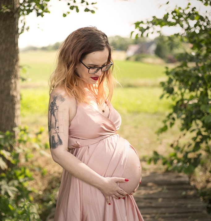 pregnant woman photo with baby pink dress holding her belly in front of park scenery